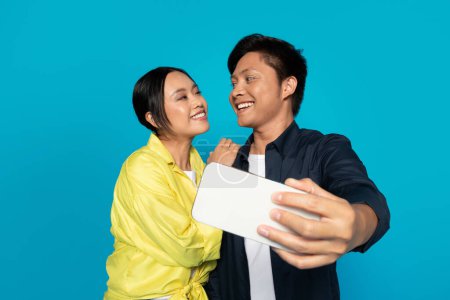 Photo for Cheerful asian couple in casual make selfie at smartphone, rejoicing date, isolated on blue studio background. App for online blog, video call and photo for social networks - Royalty Free Image