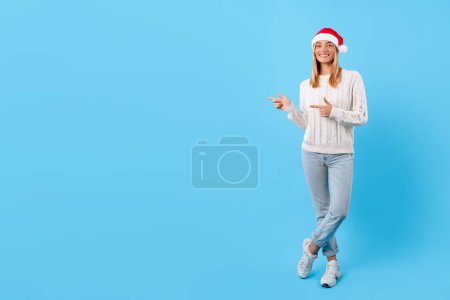 Photo for Joyful young lady wearing Santa hat and white sweater casually pointing to the side at free space with smile on bright blue background, suggesting product - Royalty Free Image