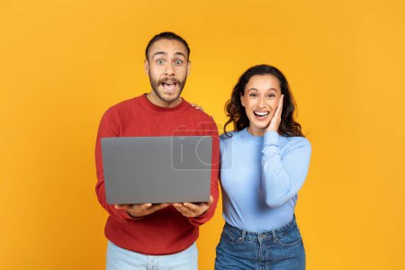 Photo for Amazing offer, good site. Surprised millennial husband and wife holding laptop and looking at camera with open mouth, posing on yellow background, studio shot - Royalty Free Image