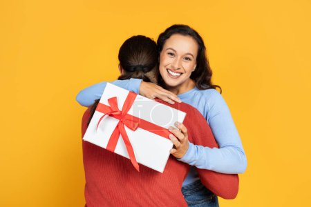 Photo for Loving man making birthday surprise for his girlfriend, happy young lady with gift box in her hand holding gift box, celebrating anniversary together, yellow studio background - Royalty Free Image