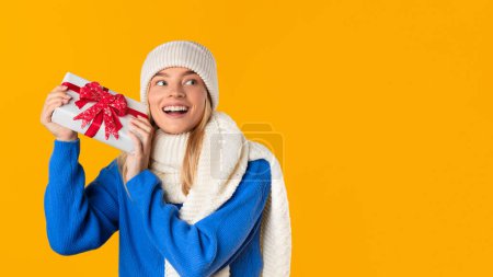 Téléchargez les photos : Joyeuse jeune femme en tenue d'hiver tient excitamment cadeau de Noël, affichant bonheur et anticipation sur fond jaune vif - en image libre de droit