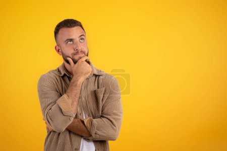 Téléchargez les photos : Focalisé jeune homme caucasien coûteux avec barbe dans des pensées décontractées, isolé sur fond de studio orange. Choix, mode de vie et émotions humaines, publicité et offre de travail, vente, création d'idées - en image libre de droit
