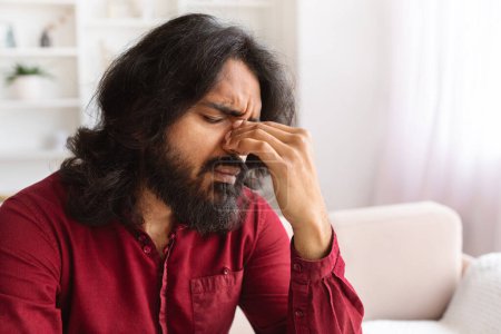 Photo for Unhappy tired handsome bearded indian young man in smart casual experiencing problems in life, sitting alone on couch at home, touching his nose bridge, suffering from migraine, copy space - Royalty Free Image