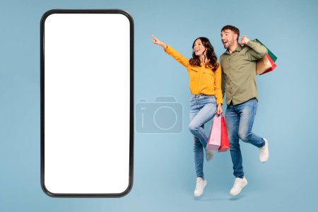 Foto de Jóvenes cónyuges europeos saltando con bolsas de compras, mujer apunta a un teléfono celular gigante con pantalla en blanco, perfecto para anuncios de ventas, sobre fondo de estudio azul - Imagen libre de derechos