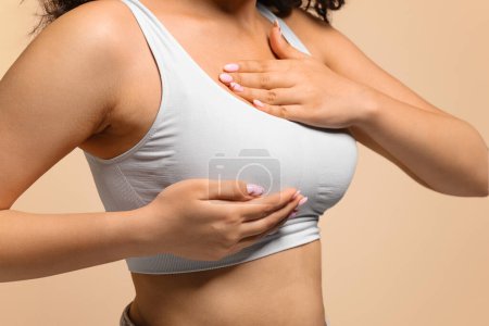 Téléchargez les photos : Jeune femme méconnaissable dans le soutien-gorge blanc main supérieure vérifiant des grumeaux sur son sein, Millennial femelle examinant le corps pour des signes de cancer du sein, debout isolé sur fond beige, recadré - en image libre de droit