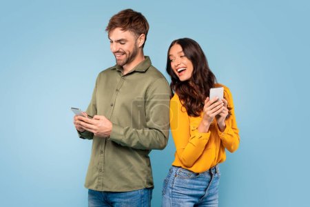 Photo for Curious young European woman subtly glancing at her husbands cellphone, both smiling against blue studio background, emanating interest and suspicion - Royalty Free Image