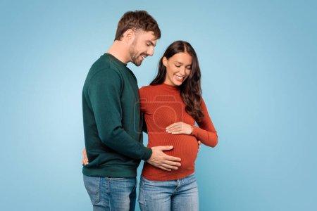 Foto de Joven europeo abraza calurosamente a su esposa embarazada, sus manos amorosamente colocadas sobre su vientre mientras sonríen juntas contra el fondo azul del estudio - Imagen libre de derechos