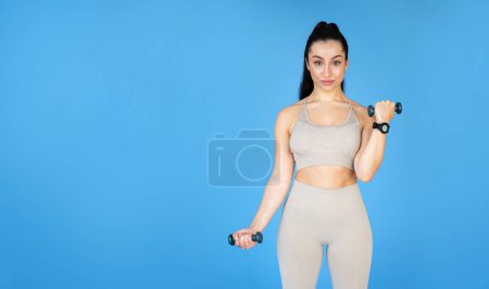 Foto de Una joven mujer en forma está haciendo ejercicios de fuerza con pesas frente a una pared azul. Ella en ropa de ejercicio, mostrando su energía y aliento, espacio vacío para el anuncio junto a ella - Imagen libre de derechos