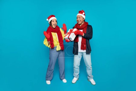 Téléchargez les photos : Heureux jeune homme coréen en chapeau donner cadeau à la femme choquée, profiter des vacances d'hiver, cadeaux de Noël, isolé sur fond de studio bleu. Nouvel An surprise, célébration ensemble - en image libre de droit