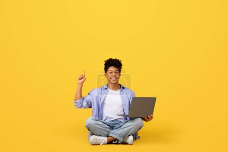 Téléchargez les photos : Étudiant noir élégant en studio utilise un ordinateur portable et pointe vers le haut à l'espace vide au-dessus de lui sur fond jaune vif, symbolisant l'innovation et les idées - en image libre de droit