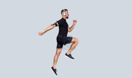 Photo for Focused young caucasian guy in wireless headphones, sportswear listen music, run, jump, freeze in air, enjoy body care and cardio workout, isolated on gray studio background - Royalty Free Image