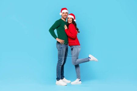 Foto de Cónyuges europeos felices en sombreros de Santa abrazándose de pie sobre fondo azul, celebrando Año Nuevo o Navidad juntos. Longitud completa de pareja joven en traje festivo posando en el estudio - Imagen libre de derechos