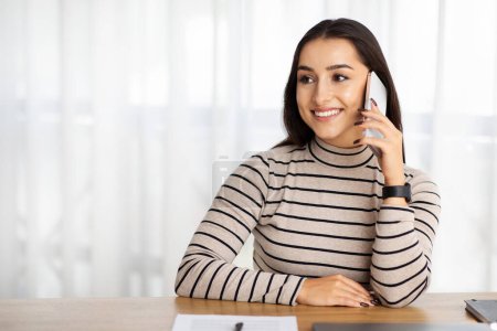 Photo for Cheerful pretty millennial arab lady manager work, call by smartphone at table, talk in living room interior. Conversation, gossip and communication with client at home, good news at free time - Royalty Free Image