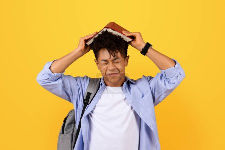 Photo for Overwhelmed young black man in casual outfit with backpack places red book on his head, eyes closed, showing signs of stress or frustration against yellow background - Royalty Free Image