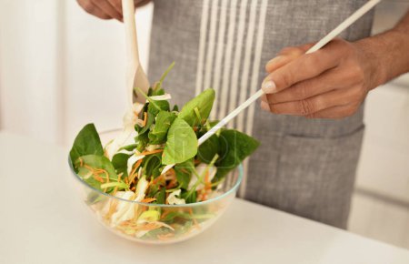 Téléchargez les photos : Un noir méconnaissable mélange des ingrédients de légumes frais pour préparer une salade pour le déjeuner, prépare un plat végétarien nutritif dans la cuisine moderne de la maison, plan recadré. Bien-être et nutrition - en image libre de droit