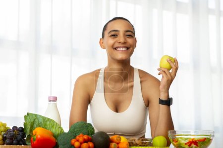 Photo for Cheerful millennial latin lady in sportswear eat green apple in light kitchen interior, enjoy fresh vegetables, salad. Diet, weight loss, healthy food, vegan and vitamins at home - Royalty Free Image