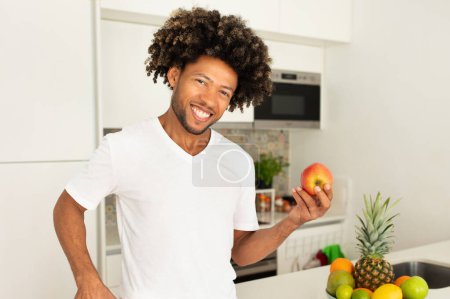 Photo for African American guy smiling, holding apple in modern kitchen, embodying healthy lifestyle and nutritious choices. Happy fit man posing with fruit, recommends wellbeing and weight loss - Royalty Free Image