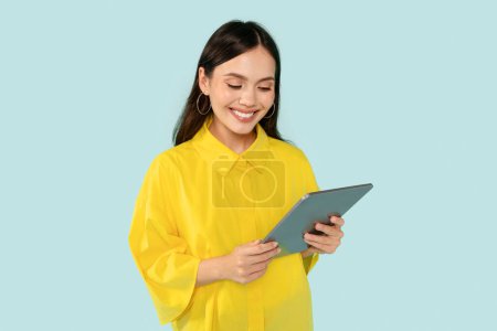 Photo for Cheerful pretty young woman wearing yellow shirt posing isolated on blue studio background, using modern pc digital tablet, checking nice educational or entertaining mobile app, copy space - Royalty Free Image
