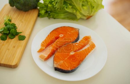 Photo for Plate with two fresh slices of salmon rich in Omega 3 lie on wooden board on kitchen counter tabletop near green vegetables indoor, healthy culinary preparation for weight loss meal - Royalty Free Image