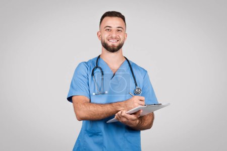 Téléchargez les photos : Infirmière masculine joyeuse en blouse bleue tenant un presse-papiers et un stylo avec stéthoscope autour du cou, symbolisant la documentation sur les soins de santé et les soins aux patients - en image libre de droit