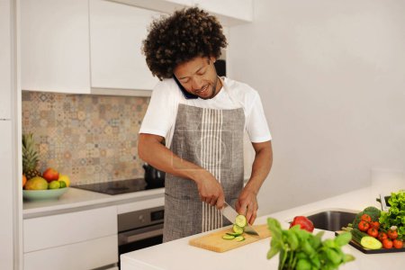 Téléchargez les photos : Jeune noir en tablier parle sur son téléphone portable tout en préparant un déjeuner sain, couper des légumes frais pour la salade à l'intérieur de la cuisine de la maison, mettant en valeur la synergie entre l'alimentation et le bien-être - en image libre de droit