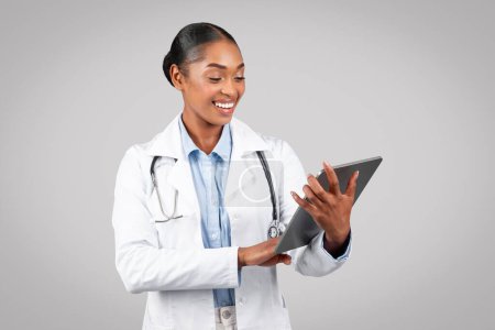 Foto de Sonriendo bastante millennial terapeuta médico mujer negro en blanco capa escribiendo en la tableta, charlando, aislado en el estudio de fondo gris. Aplicación de atención médica de forma remota, consulta - Imagen libre de derechos