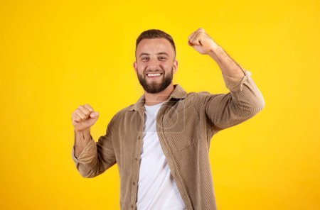 Photo for Happy millennial european man with beard in casual has fun, rise fists, celebrate win, isolated on yellow studio background. Victory, success, lifestyle and human emotions, ad and offer - Royalty Free Image