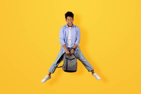 Foto de Chico negro alegre en salto de aire juguetón, sosteniendo su mochila con amplia sonrisa, vestido con atuendo casual contra fondo amarillo brillante, longitud completa - Imagen libre de derechos