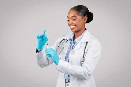 Photo for Cheerful millennial black lady doctor in white coat, protective gloves use syringe, isolated on gray background studio. Vaccination recommendation, medicine and injection, health care in clinic - Royalty Free Image