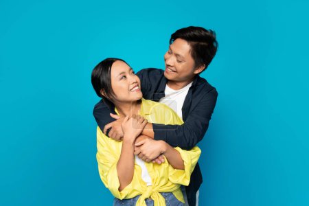 Photo for Smiling millennial korean guy hug lady in casual look at each other, enjoy relationships, isolated on blue studio background. Romantic fun, date and love, sale, ad and offer - Royalty Free Image
