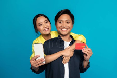Foto de Sonriente joven mujer japonesa abrazo hombre en el teléfono inteligente show casual, tarjeta de crédito, aislado en fondo de estudio azul. gadget Recomendación, banca financiera, venta en línea, aplicación de compras - Imagen libre de derechos