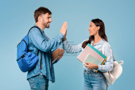 Photo for Young European students, man and woman with books and backpacks, share high five against vibrant blue studio background, embodying friendship and success - Royalty Free Image