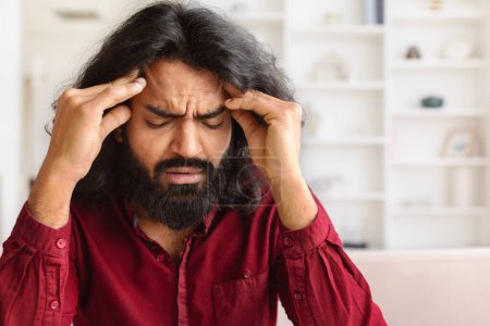 Photo for Closeup of exhausted bearded millennial indian man suffering from headache, sitting on couch with closed eyes, touching his head, home interior, copy space - Royalty Free Image