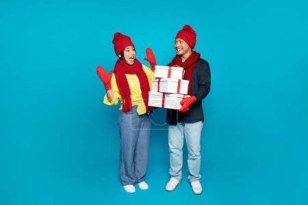 Téléchargez les photos : Heureux jeune chinois dans le chapeau donne de nombreuses boîtes présentes pour surprendre femme, isolé sur fond de studio bleu, pleine longueur. Couple profiter des vacances d'hiver et du Nouvel An, cadeaux de Noël - en image libre de droit