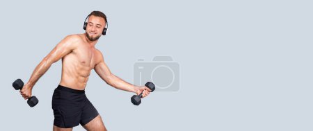 Téléchargez les photos : Joyeux musclé millénial homme caucasien athlète avec torse nu dans les écouteurs sans fil, écouter de la musique, faire des exercices avec haltère, isolé sur fond de studio gris, panorama, annonce, offre - en image libre de droit
