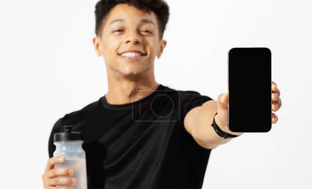 Photo for Brazilian fitness guy recommends workout app on smartphone, showing black phone screen and holding water bottle on white studio background. Cropped, mockup for gym advertisement - Royalty Free Image