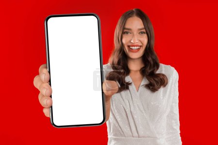 Photo for Joyful millennial woman holds big smartphone with blank screen in red studio, dressed in festive attire. Mockup for mobile holiday themed designs, Valentines Day and birthdays. Collage - Royalty Free Image