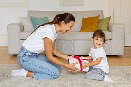 Photo for Childs Birthday. Loving Mother Giving Gift To Little Daughter While Sitting Together On Floor At Home, Mommy Congratulating Toddler Girl With Holiday, Surprising With Present, Free Space - Royalty Free Image