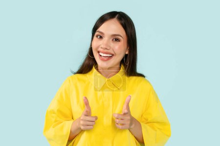 Téléchargez les photos : Positif joyeux attrayant jeune femme portant chemise jaune indiquant pointant vers la caméra et souriant à la caméra, jolie millénaire dame flirter, posant sur fond bleu studio - en image libre de droit