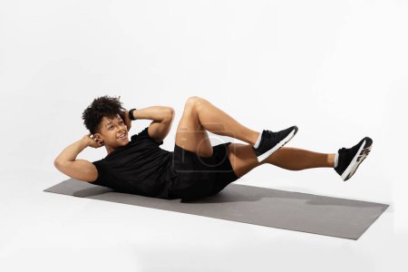 Photo for Athletic young man does elbow to knee crunches on fitness mat, working on his abdominal muscles and core strength, lying on white studio backdrop. Workout for perfect abs concept - Royalty Free Image