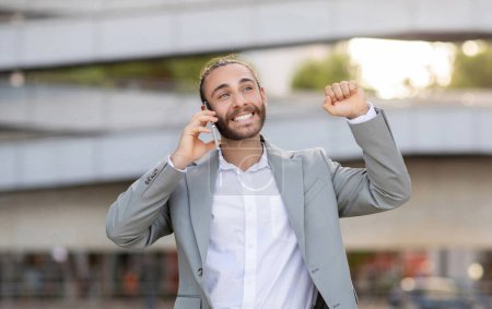 Photo for Joyful young businessman celebrating success during outdoor phone call, happy excited male entrepreneur having cellphone conversation with business partner, raising his hand in triumph, got good news - Royalty Free Image