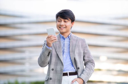 Foto de Korean Young Business Man Usando la aplicación de búsqueda por voz que sostiene el teléfono celular cerca de la boca y hablando, utilizando el servicio de asistente móvil de pie al aire libre en el área urbana de la ciudad. Comunicación en línea - Imagen libre de derechos
