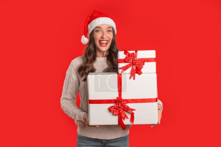 Téléchargez les photos : Dame souriante au chapeau de Père Noël tient deux boîtes-cadeaux enveloppées dans un cadre de studio rouge. Bannière avec femme en tenue de fête avec cadeaux pour Noël et Nouvel An Publicité - en image libre de droit