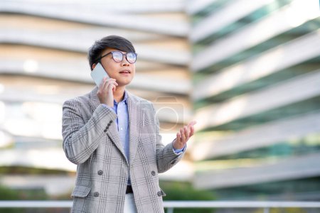 Photo for Young Asian specialist man talking via smartphone in hand, communicates new digital business strategy on urban street with office buildings outside, free space for text advertisement - Royalty Free Image