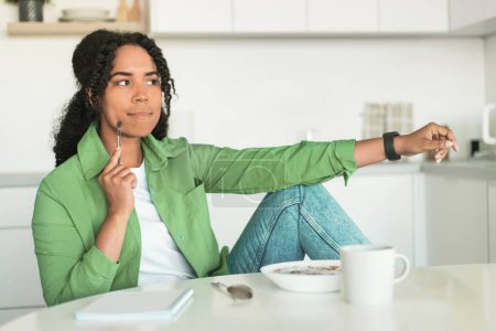 Photo for Pensive black woman taking notes sitting at her breakfast, holding pen and thinking about diet plan, learning in modern kitchen interior, looking aside. Young lady plans her day, writing tasks - Royalty Free Image