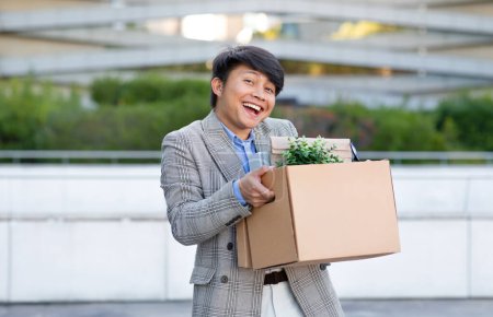 Téléchargez les photos : Joyeux homme d'affaires asiatique avec boîte à effets célébrant le nouveau début de carrière, se tient à l'extérieur près de l'immeuble de bureaux moderne, exsudant l'optimisme de la liberté et des possibilités d'emploi - en image libre de droit