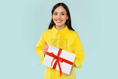 Photo for Positive beautiful brunette young woman wearing yellow shirt holding white gift box with red bow and smiling at camera, going to birthday party, posing isolated on blue background - Royalty Free Image