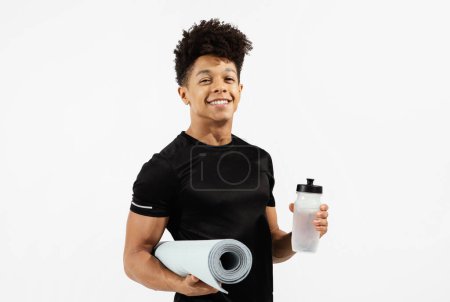 Photo for Athletic young man holds water bottle and fitness mat, hydrating during workout, standing in black t-shirt on white studio backdrop, embodying health and strength. Sporty lifestyle - Royalty Free Image