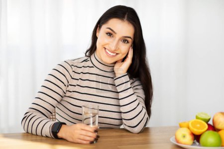 Foto de Sonriente mujer árabe joven beber vaso de agua en la sala de estar, interior de la cocina en la mesa con frutas. Señora disfrutar de vitaminas, atención médica y desayuno, dieta y estilo de vida en el hogar - Imagen libre de derechos