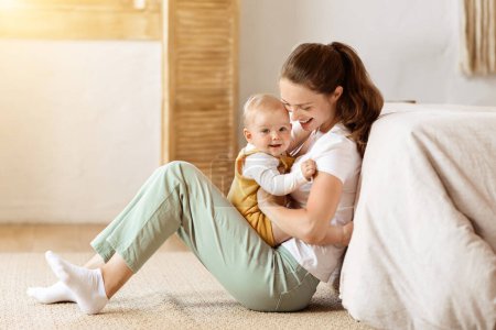 Photo for Young Mom Posing With Baby Toddler Holding Helping Her Son To Sit. Happy Mother Caring For Child Infant Bonding And Playing With Cute Little Boy, Bedroom Interior. Copy Space - Royalty Free Image